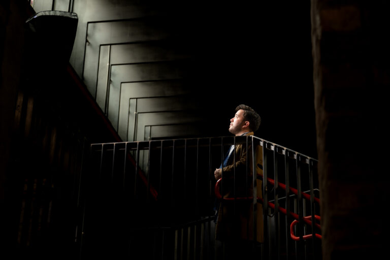 Aage standing in a stairway looking up to the windows where the light is coming from.